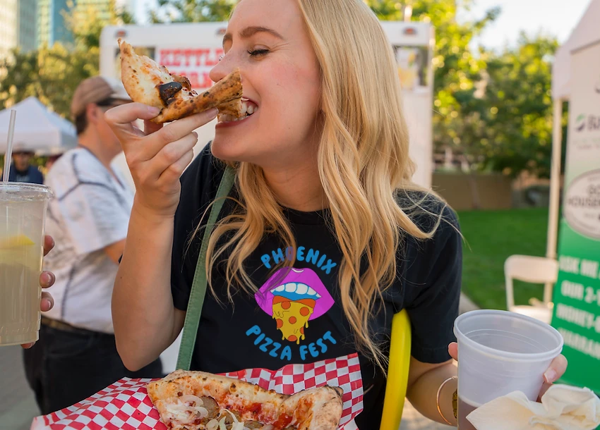 Woman in a Phoenix Pizza Fest tee biting a slice at the festival