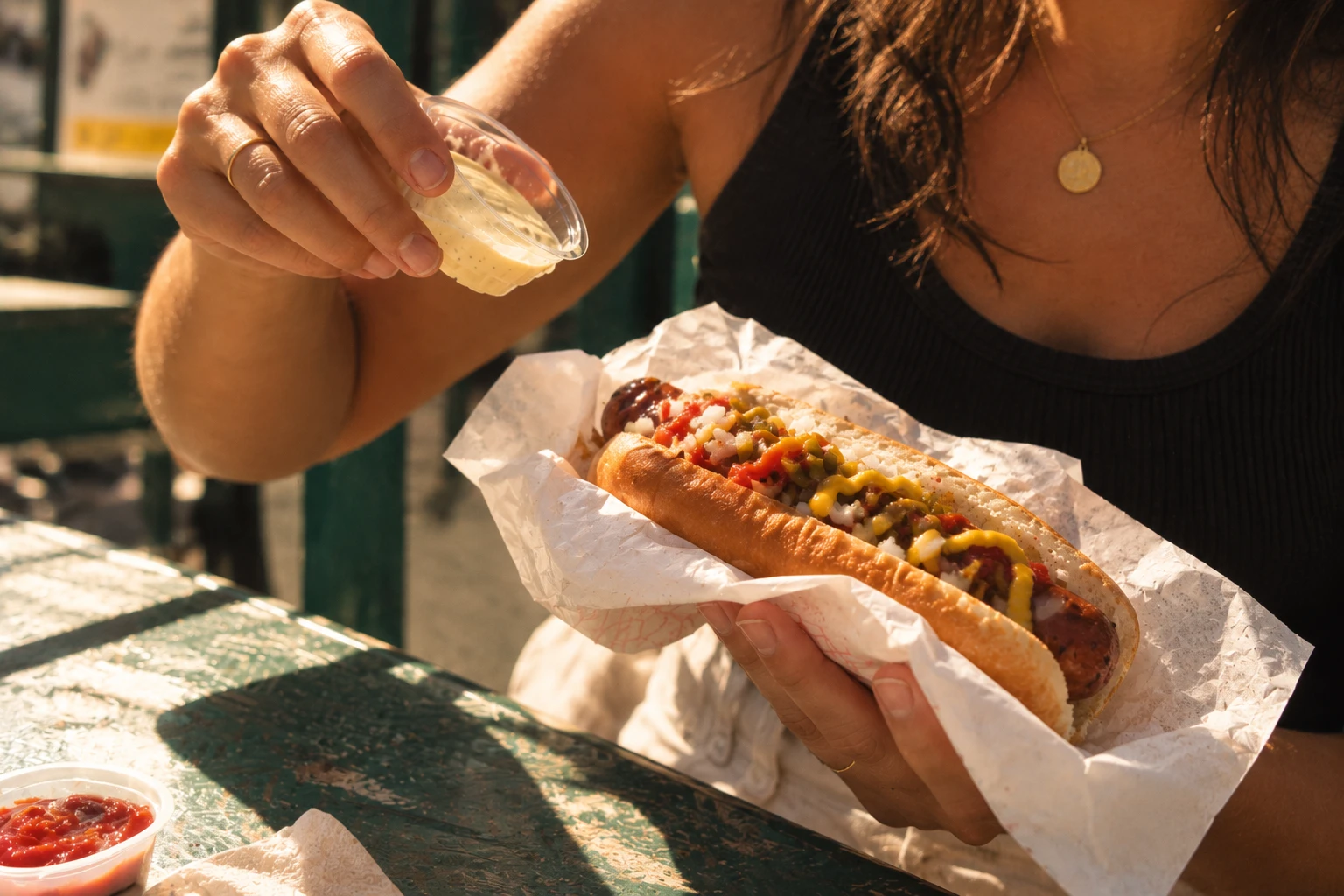 Hands adding sauce to a loaded Wurst hot dog at a sunny green table